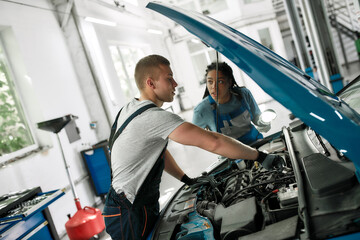 Obraz premium Apprenticeship. Professional male mechanic repairing car engine, tighten, screw with spanner while his female colleague looking at him, holding torch under car hood at service station