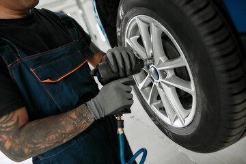 Cropped shot of male mechanic screwing or unscrewing car wheel of lifted automobile by pneumatic wrench at auto repair shop