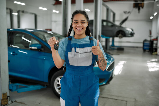 Best service here. Young african american woman, professional female mechanic smiling at camera, giving car key and showing thumbs up in auto repair shop