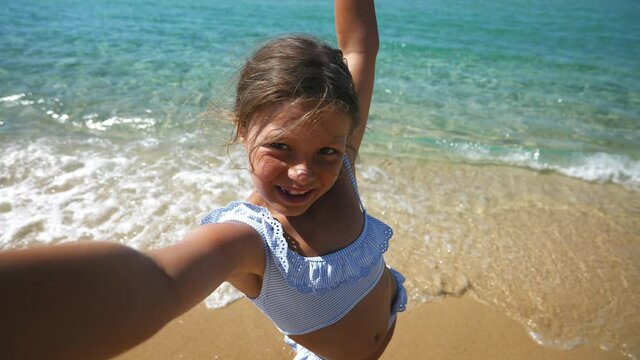 A happy carefree little girl is having fun to make a selfie or technology video call to parents or friends on a beach with seaside in a sunny day during family vacation holidays.