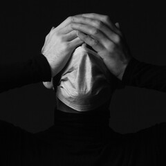 Coronavirus, Bio Protection Concept. Close up portrait of crying man wearing medical mask, black turtleneck, hiding face with hands. Monochrome studio shot