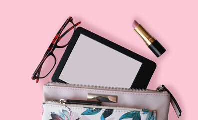 top view of a purse with an ebook reading glasses and lipstick.  Pink pouch flatlay with an...