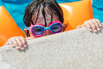 Little black-haired girl in the pool wearing sleeves and swimming goggles, peeking over the curb