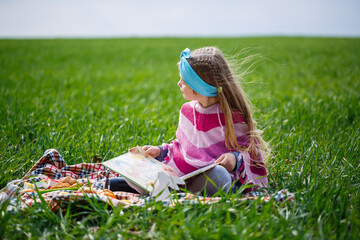 little girl child sits on a bedspread and reads a book with a fairy tale, green grass in the field, sunny spring weather, smile and joy of the child, blue sky with clouds