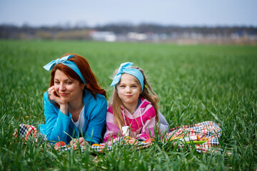 little girl child and mother woman lie on the bedspread, green grass in the field, sunny spring weather, smile and joy of the child, blue sky with clouds