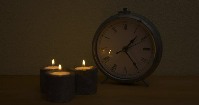 Traditional clock with roman numbers at nighttime on wooden table with three lit candles