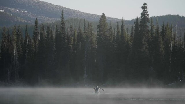 girl person with dog on kayak paddle across misty fog covered water lake with forest trees and mountains background peaceful morning sparks lake central oregon usa slow motion