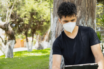 Hispanic young man with mask under the tree using his digital tablet in the park on a sunny day - young man sitting on the grass studying with electronic device - social distancing