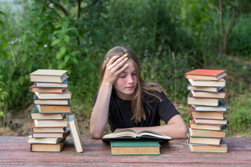 Young girl reading a book in the garden at a wooden table with a stack of books