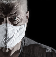 Portrait of an older man with gray hair. He is wearing a medical mask. The background is black.