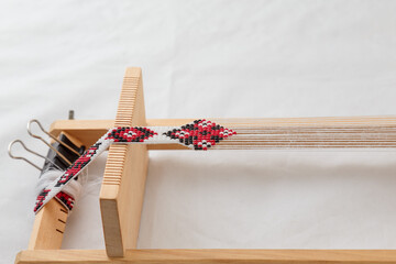Wooden beadwork machine with strained white threads and part of ready beaded necklace with ethnic national ornament. Making handmade jewelry, female bijouterie, hobby and leisure concept.