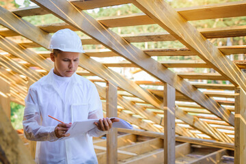 Naklejka na ściany i meble Foreman holding drawings, looking the project of wooden house, working on a construction site. Young attractive engineer in a white helmet checks the roof frame. Concept career architect. Copy space.