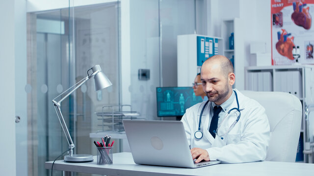 Doctor typing an e-mail from modern office of private clinic with glass walls, nurse in background checking X Rays, medical staff and patients walking in busy hallway. Healthcare system clinic