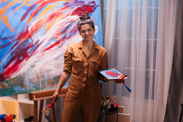 Happy young painter holding wooden palette for paint in art workshop. Modern artwork paint on...