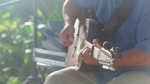 Man plays twelve string acoustic guitar during an outdoor party in pretty backlit sunlight. 