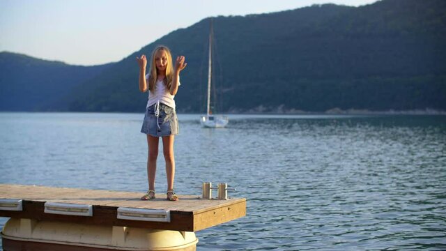 Happy girl teenager dancing on pier on sea lake green hills and yacht landscape. Cheerful young girl dancing on sea pier on green mountains background at summer vacation