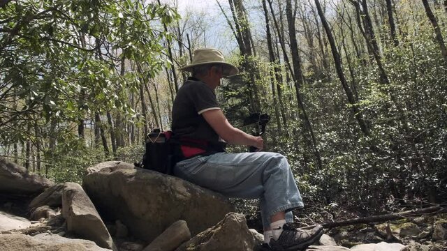 Adult Caucasian woman hiker sits alone on outdoor rock takes video of natural green forest landscape with mobile phone and gimbal on sunny day, handheld low angle close up profile