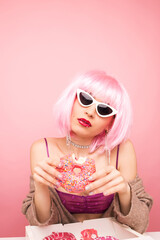 Stylish girl in a pink wig eats delicious donuts. Shot in a studio on a pink background.