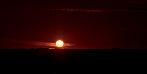 The sun setting in the ocean with small islands and a lighthouse in the horizon.