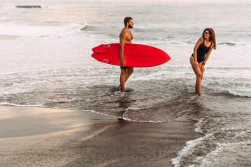 Fototapeta na wymiar Beautiful couple have fun at the sea. Couple in love on the beach with a surfboard at sunset. Beautiful couple on the island of Bali. Couple of surfers walking on coast in Indonesia. Vacation at sea