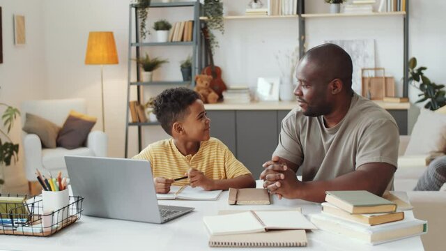 African American father sitting with little son at table in the living room, pointing on laptop screen and explaining homework to kid as he taking notes