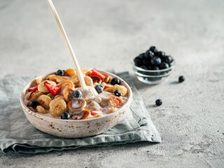 Trendy food - pancake cereal. Milk pouring on heap of mini cereal pancakes in bowl. Tiny cereal pancakes with berries in craft plate over gray cement background. Copy space for text or design.