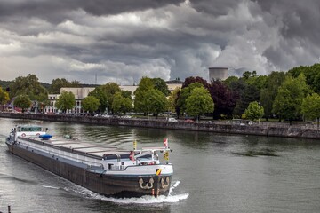 Cargo barge sails along the river Maas. Green quay and dramatic cloudy sky on the background.