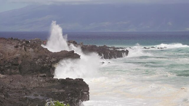 Large wave crashes on rocky Hawaiian coastline in Maui. Rough dangerous surf fills the coast as tourists take photos in the distance. Molokai in the background. 