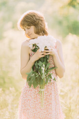 Beautiful young Caucasian blond woman in light dress holding bouquet of white peonies, walking in summer field or garden in sunset. Woman with flowers outdoors