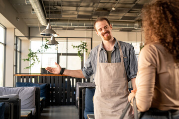 Smiling handsome young waiter in apron gesturing to free table while welcoming girl in cozy cafe