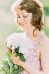 Naklejka premium Beautiful young Caucasian blond woman in light dress holding bouquet of white peonies, walking in summer field or garden in sunset. Woman with flowers outdoors