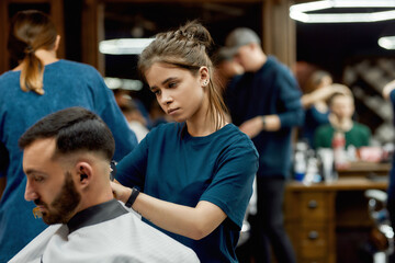 Obraz premium Focused professional barber girl making trendy haircut for a young bearded man sitting in barber shop chair. Side view. Focus on a girl