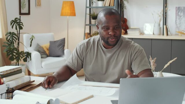 African American male architect sitting at desk in living room and discussing construction plan on online video call on laptop while working remotely at home