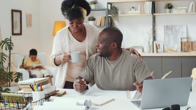 African American male architect speaking with colleague on video call on laptop while wife bringing him tea mug, giving hugs and joining conversation; little son using tablet in background