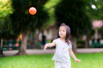 Happy Asian girl aged 3 years old throws tiny orange ball playing alone on green lawn at park in afternoon of rainy season. Sweet smile children exercise. Child is wearing white shirt and long pants.