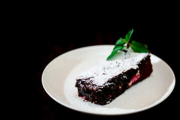 Chocolate brownie piece on white plate decorated with mint leaves and sugar powder on black background