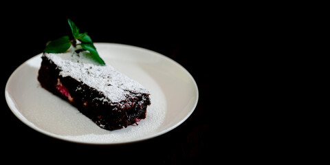 Chocolate brownie piece on white plate decorated with mint leaves and sugar powder on black background