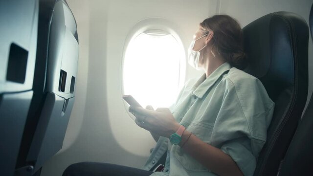 Handheld close-up shot. A young woman in a facial protective mask using her smartphone on board of an aircraft, sitting by the airplane window.