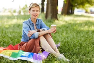 Portrait of young woman with short blonde hair smiling at camera while sitting on the grass and drinking coffee in the park