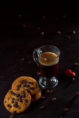 Close up of a glass cup of coffee, chocolate chip cookies and roasted coffee beans on dark background. Concept of ready to eat food, tasty snack. Selective focus, copy space.