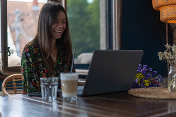 Fototapeta premium Young female using laptop, communicates on internet with customer/colleague coffee mug on table. Cozy office workplace, remote work, E learning concept