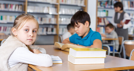 Portrait of cute preteen girl preparing for lesson in school library, reading textbooks