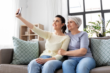 Obraz premium family, generation and people concept - happy smiling senior mother with adult daughter taking selfie by smartphone at home