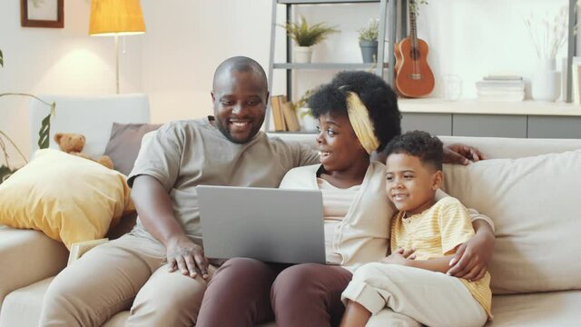 Tracking Shot Of Happy Afro-american Family Sitting Together On Couch In Living Room And Watching Movie Online On Laptop While Staying Together At Home