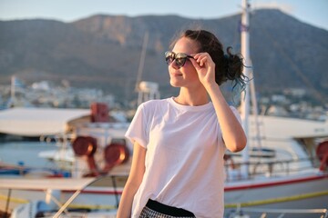 Beautiful fashionable girl teenager walking along sea promenade, pier with yachts