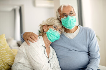 Pensive senior couple with protective face masks sitting embraced at home.
