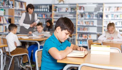Focused boy reading in school library on background with other students and teacher