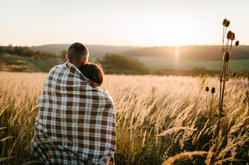 Naklejka na ściany i meble Young couple hugging, standing back, people covered with blanket, at sunset in autumn an outdoor. at field grass on background of sun. Concept of friendly family. full length. Close Up.