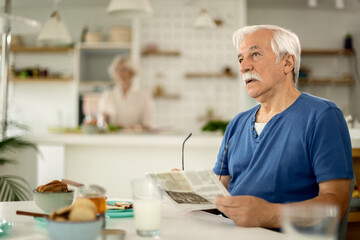 Pensive senior man reading newspaper while having breakfast at home.