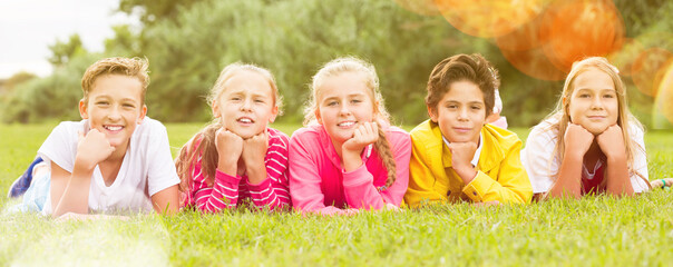 Company of five kids are posing lying on grass in the park